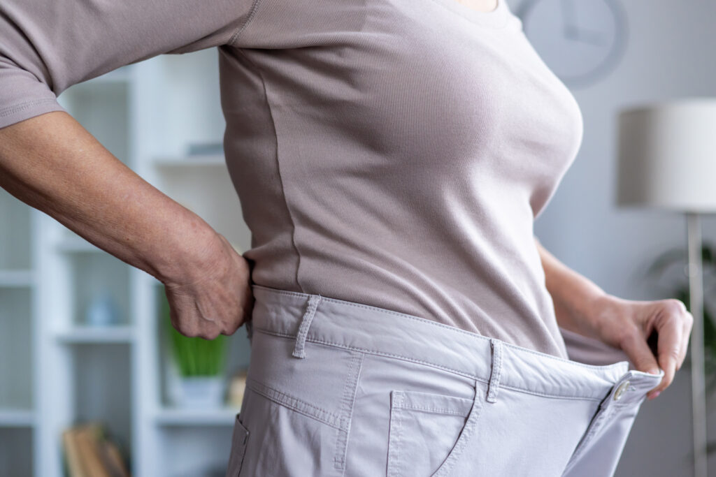 Woman shows successful weight loss progress by wearing loose pants in bright indoor setting, emphasizing healthy lifestyle transformation